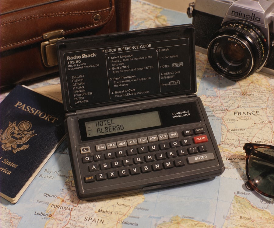 A vintage electronic translator displays “HOTEL ALBERGO” on its screen, surrounded by a camera, a US passport, sunglasses, a leather bag, and a map of Europe with “France” visible.