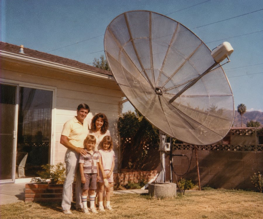A family of four stands smiling in their backyard beside a large satellite dish. The parents and two children are posing near a house with sliding glass doors and a wooden fence.