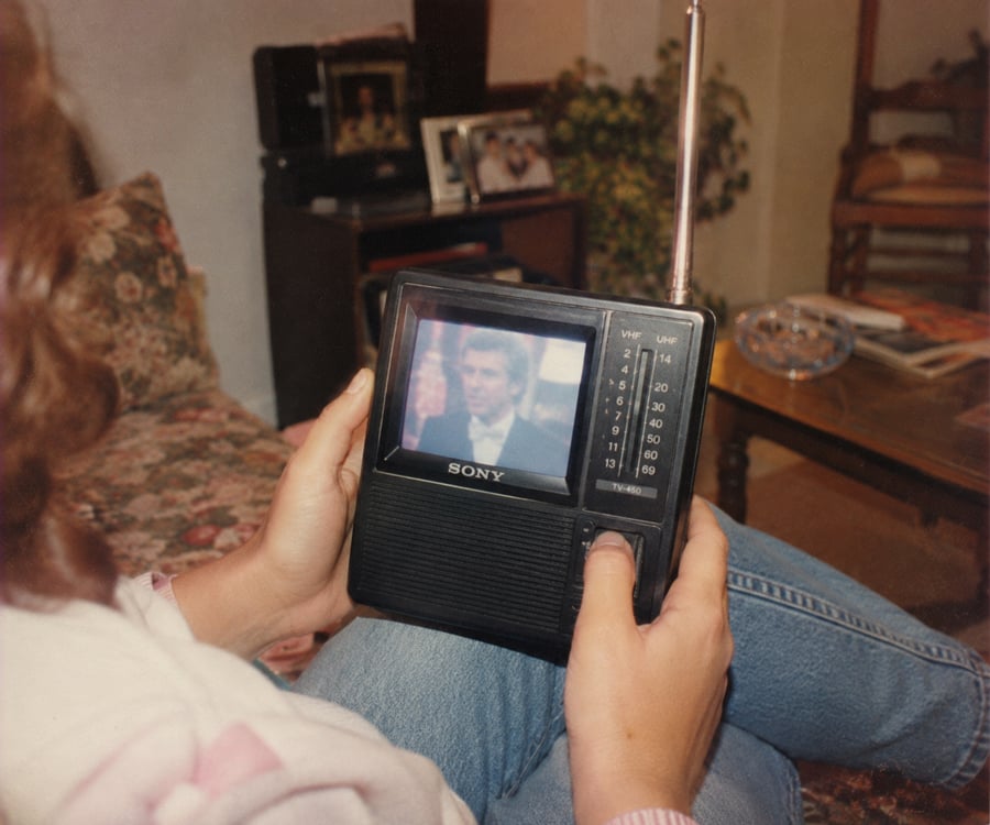 A person sits on a floral couch holding a small vintage Sony portable TV, watching a blurred image of a man on the screen. The room has plants, framed photos, and a wooden chair in the background.