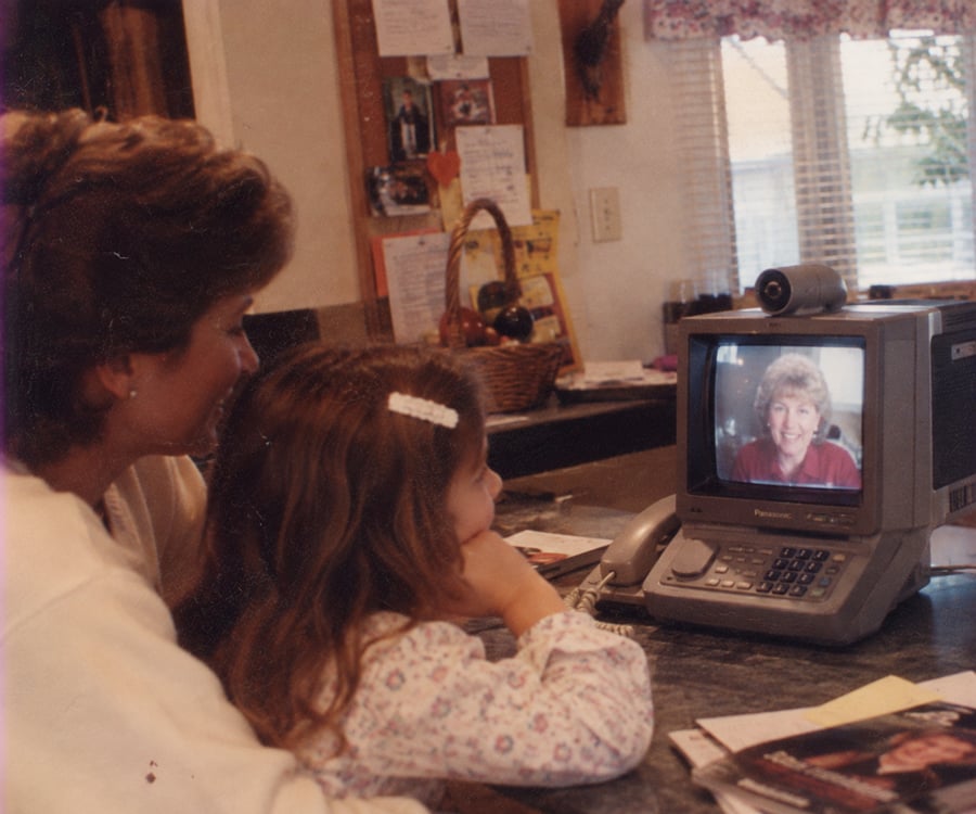 A woman and a girl sit at a kitchen counter, smiling and watching a woman on a small TV with a webcam during a video call. Papers and a phone are on the counter.