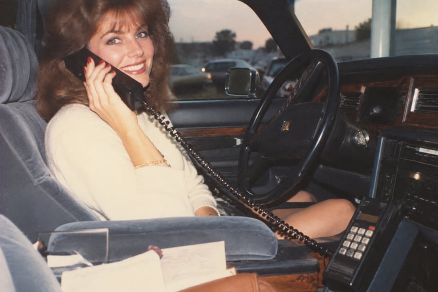 A woman with long hair smiles while sitting in a car, holding a large corded car phone to her ear. Papers and the car phone’s keypad are visible near her. The photo has a vintage, 1980s feel.