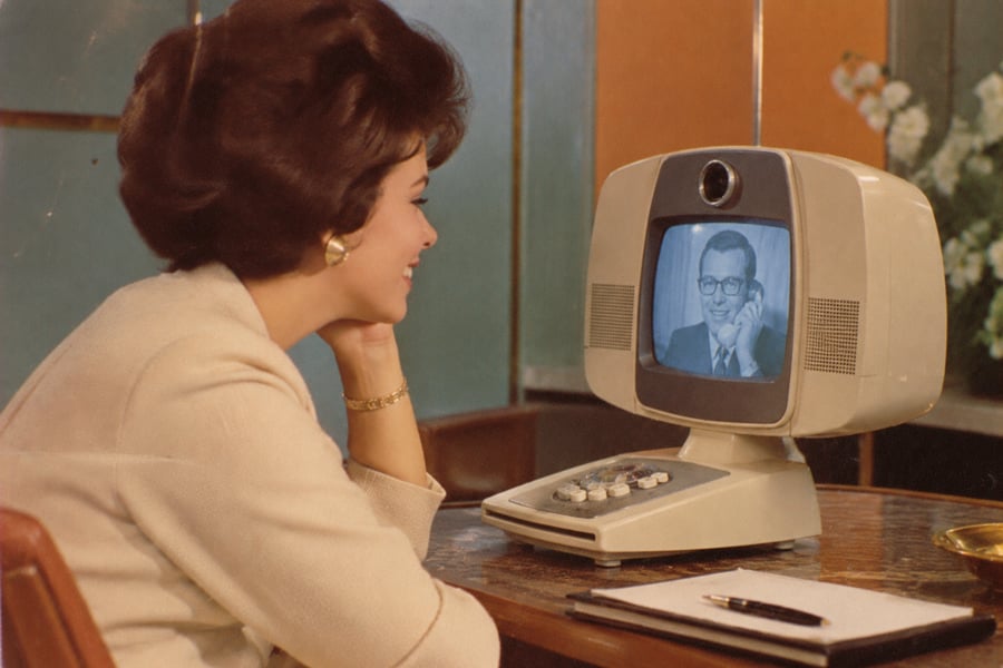 A woman sits at a desk smiling at a retro video phone displaying a man on its screen. A notepad and pen are on the desk. The background shows flowers and a stylish mid-century setting.