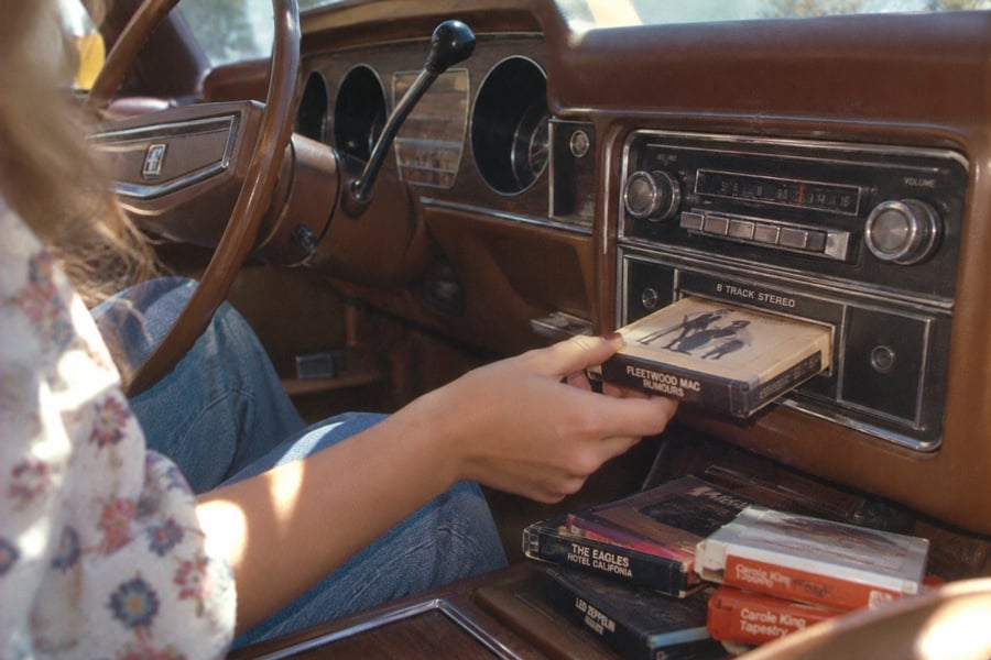 A person inserts a Fleetwood Mac 8-track tape into a vintage car stereo. Other 8-tracks, including The Eagles and Carole King, are stacked nearby on the car’s center console.