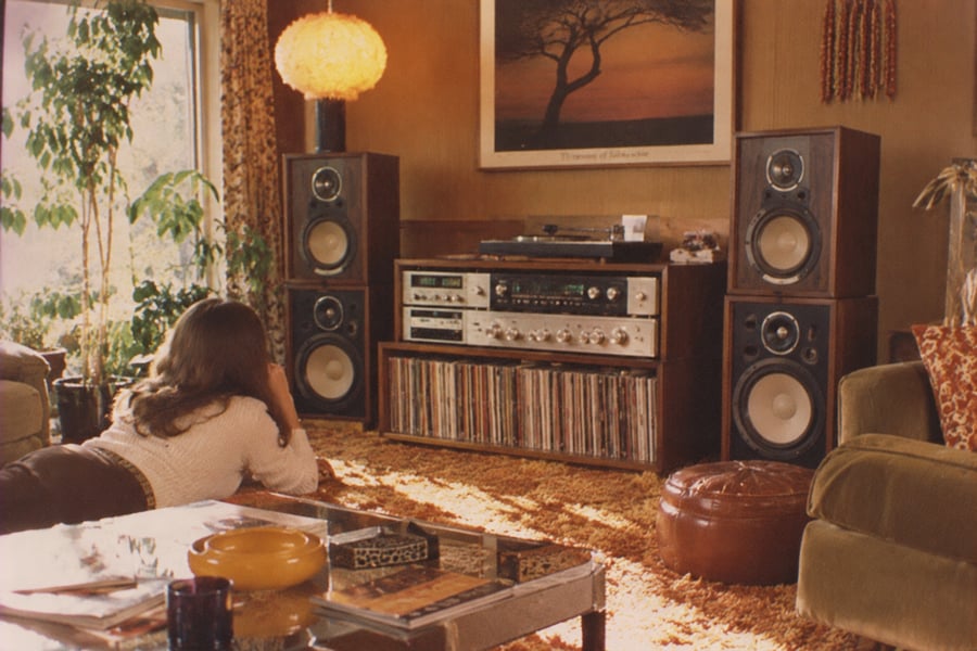 A person lies on an orange carpet in a retro living room, facing large stereo speakers and a shelf of vinyl records, with plants and a framed tree poster in the background.