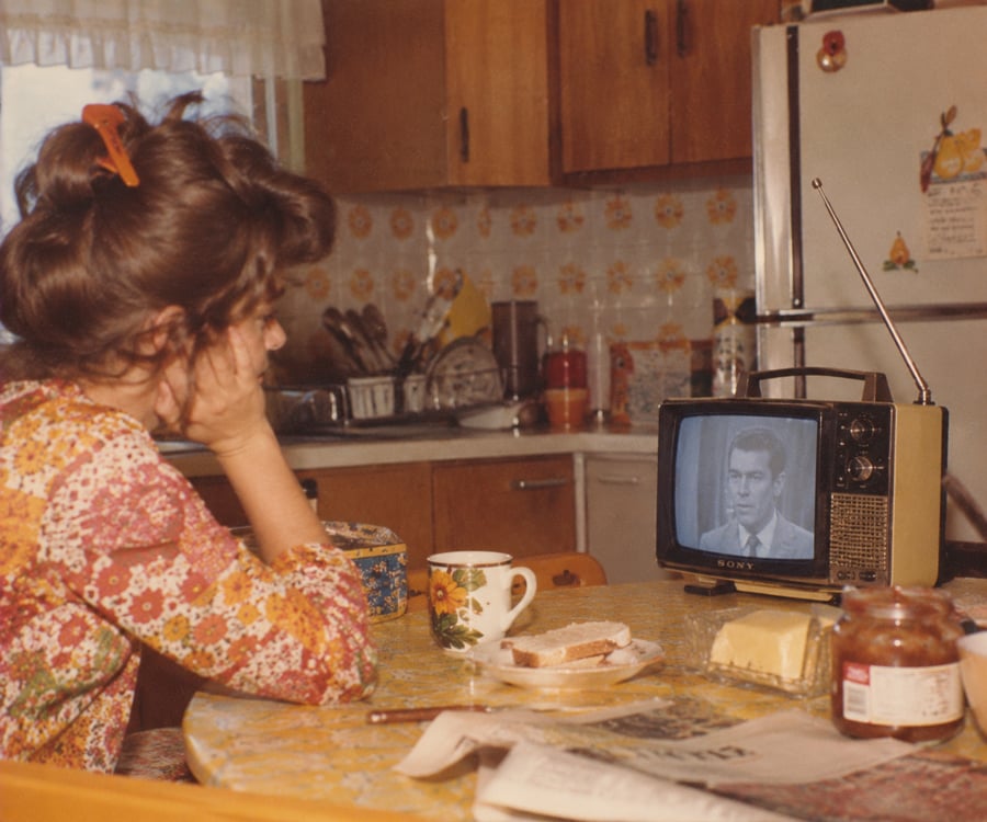 A woman in a floral dress sits at a kitchen table watching a black-and-white TV. Breakfast items, a mug, and a newspaper are on the table. The kitchen has wooden cabinets and vintage decor.