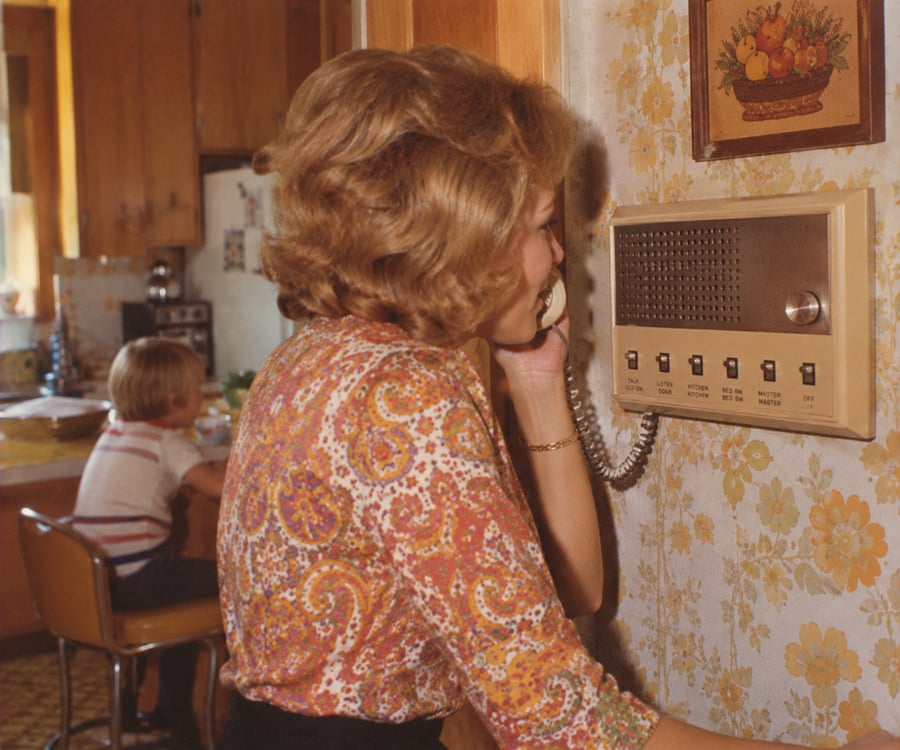 A woman with short hair uses a wall-mounted intercom phone in a vintage kitchen, while a young boy sits at a table in the background.