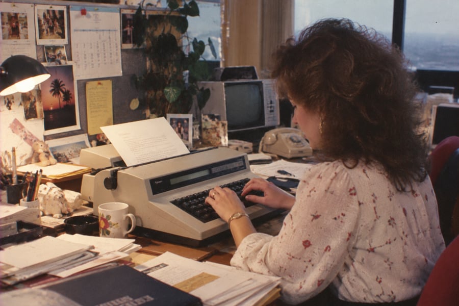 A woman with curly hair types on an electric typewriter at a cluttered office desk, surrounded by papers, a mug, telephone, and a small TV. The workspace is decorated with photos, plants, and a desk lamp.