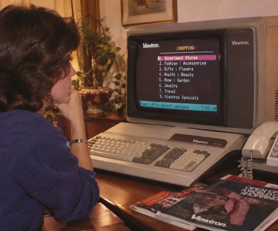 A woman sits at a desk using Viewtron, an early online shopping service, on a vintage computer. Magazines and a phone are on the desk. The screen lists shopping categories like “Department Stores” and “Fashion.”