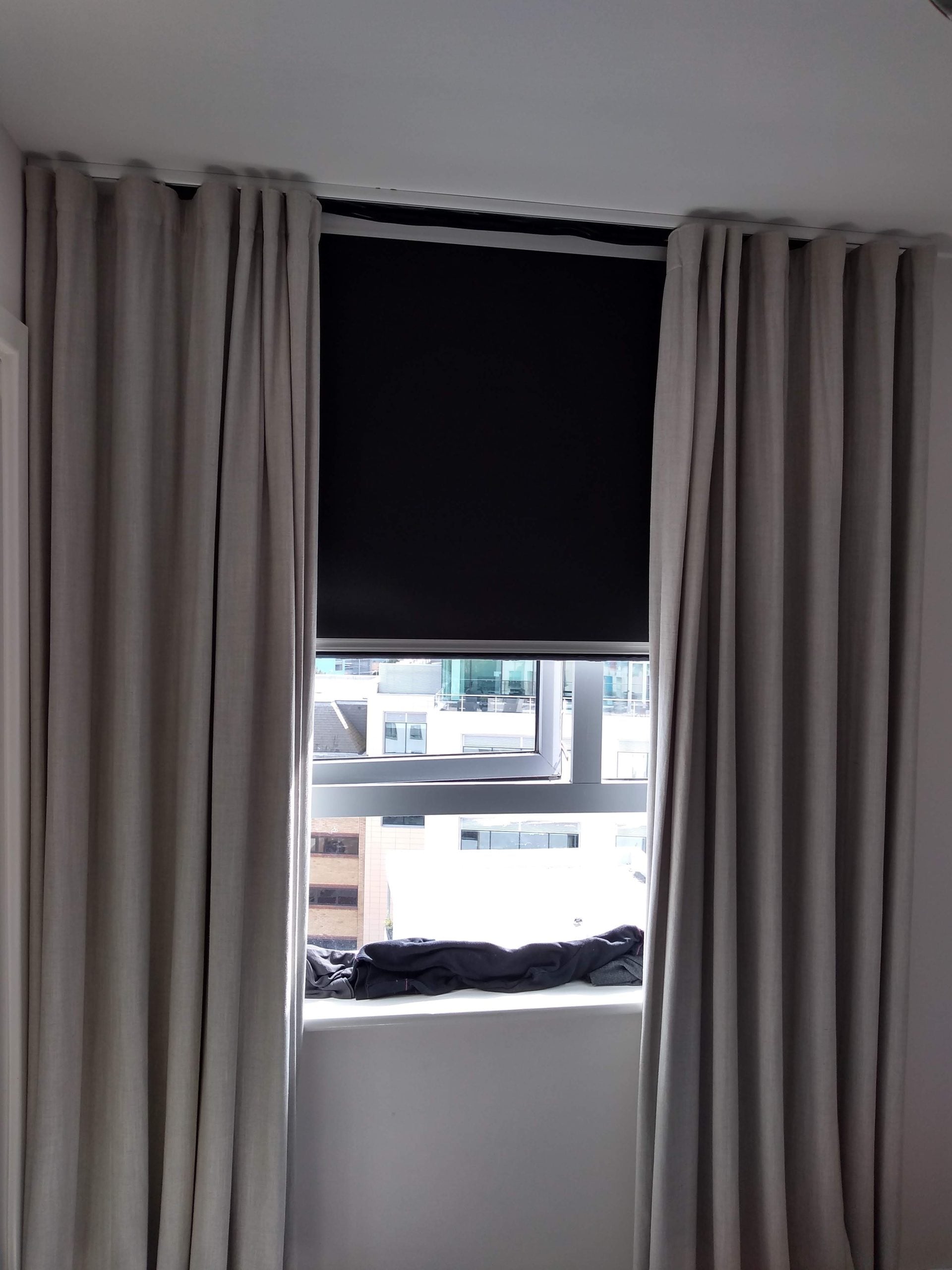 A window with beige curtains open to reveal a partially closed black roller blind. Some city buildings are visible outside through the lower part of the window.