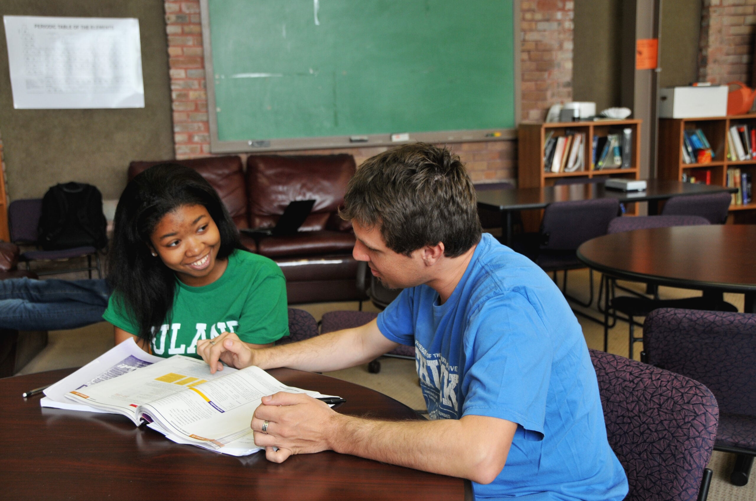 A student and a teacher sit at a table with an open textbook, having a discussion. The student smiles while the teacher points at the book. There is a chalkboard and bookshelves in the background.