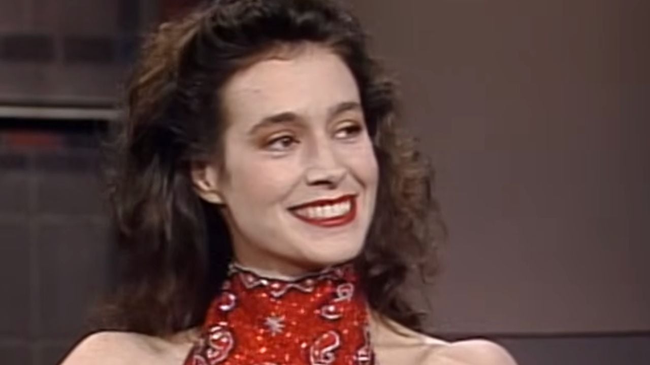 A woman with long dark curly hair smiles while wearing a red patterned halter top. The background is neutral and out of focus.