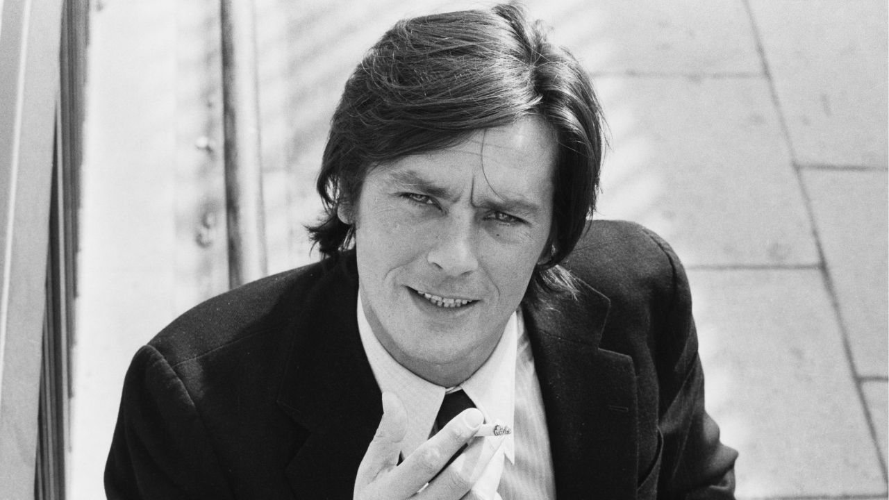 A man in a suit and tie sits outdoors, holding a cigarette and looking at the camera with a slight smile. The photo is black and white and taken from above.