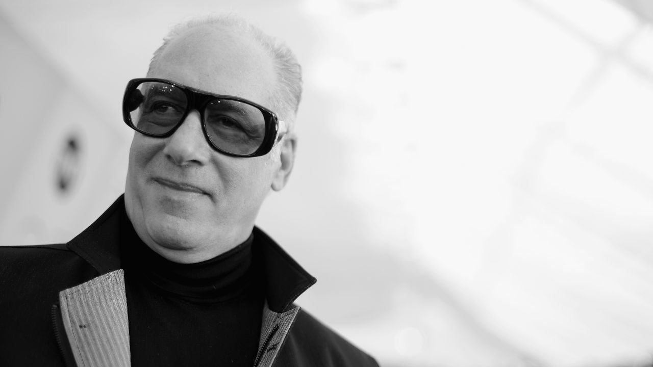 A man wearing large dark sunglasses and a high-collared jacket smiles slightly while looking away from the camera. The background is blurred and bright.