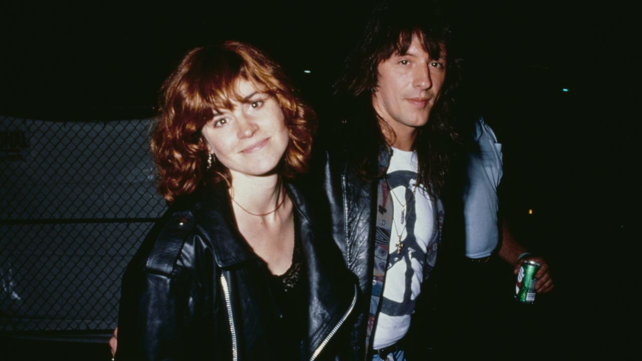 A woman and a man stand together at night, both wearing black leather jackets. The man holds a drink can, and they are near a chain-link fence. They appear to be at an outdoor event.