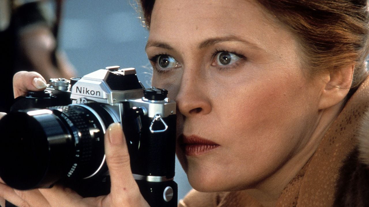 A woman with light brown hair holds a Nikon film camera close to her face, focusing intently as she prepares to take a photograph.