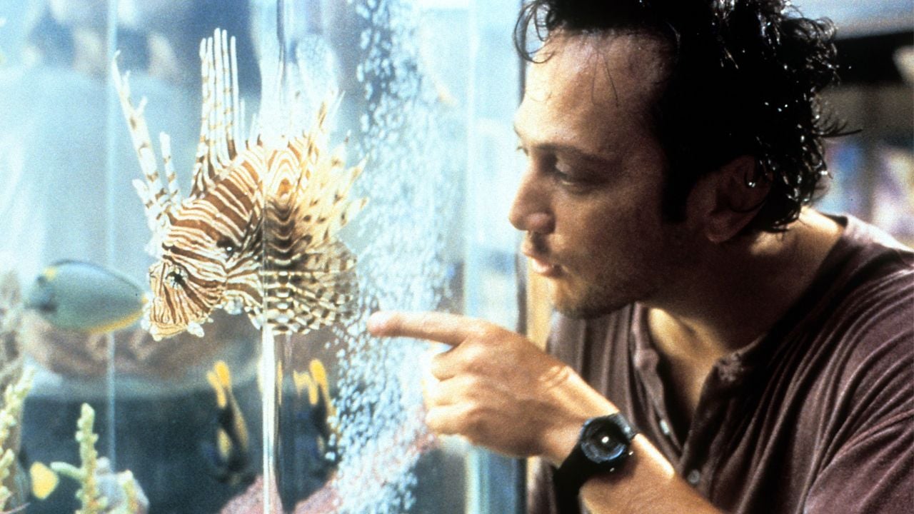 A man in a brown shirt points at a lionfish inside a large aquarium, looking closely at the fish. The aquarium has bubbles, coral, and other fish in the background.