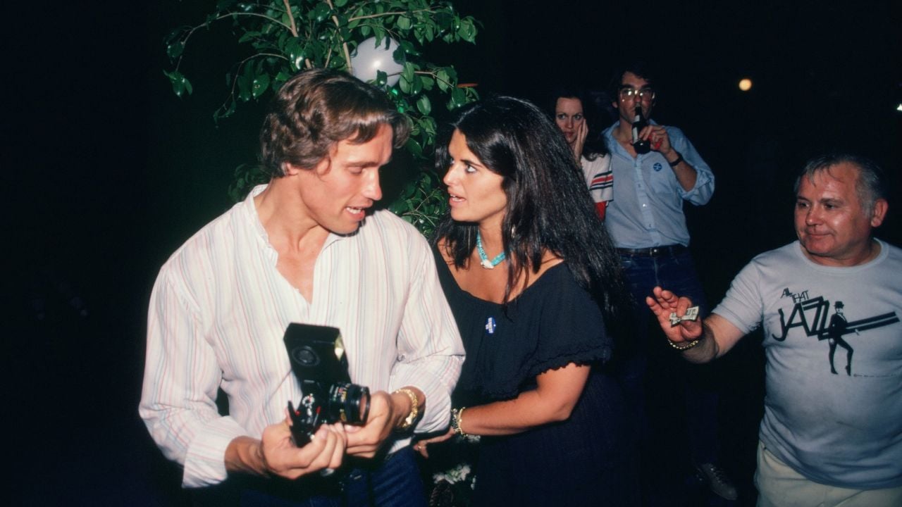 A man holding a camera talks to a woman with long dark hair, as people stand behind them in a lively, dimly lit setting with a leafy plant nearby.