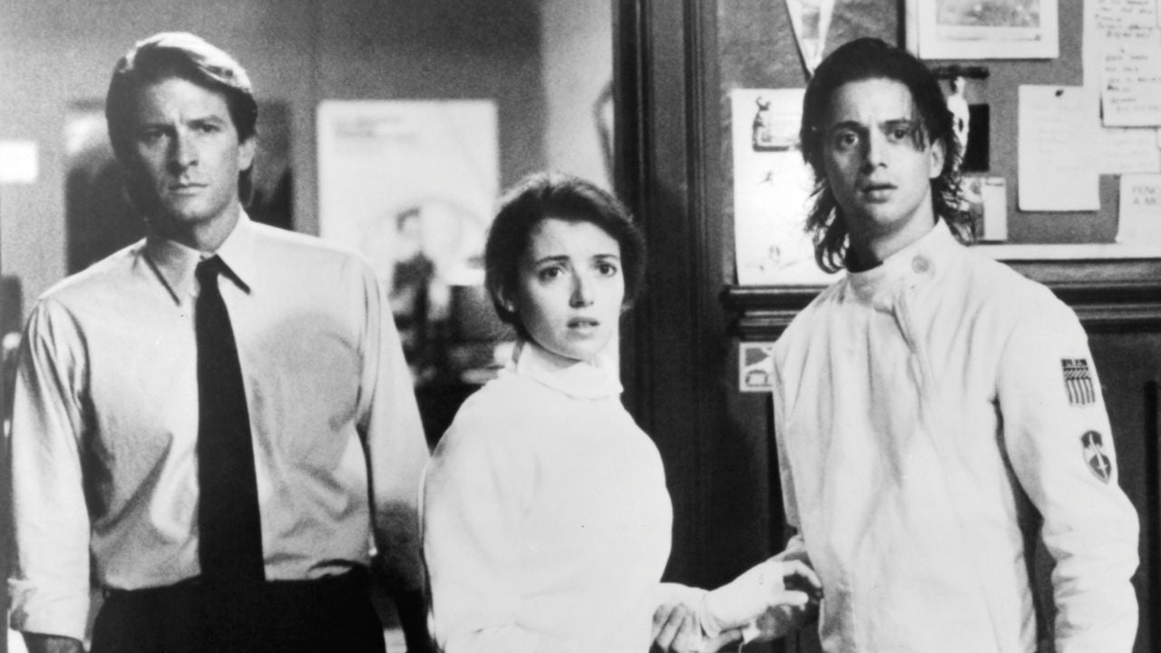 Three people stand indoors, looking surprised or concerned. One man wears a dress shirt and tie, the woman and the other man wear fencing uniforms. Bulletin boards and papers are visible on the wall behind them.