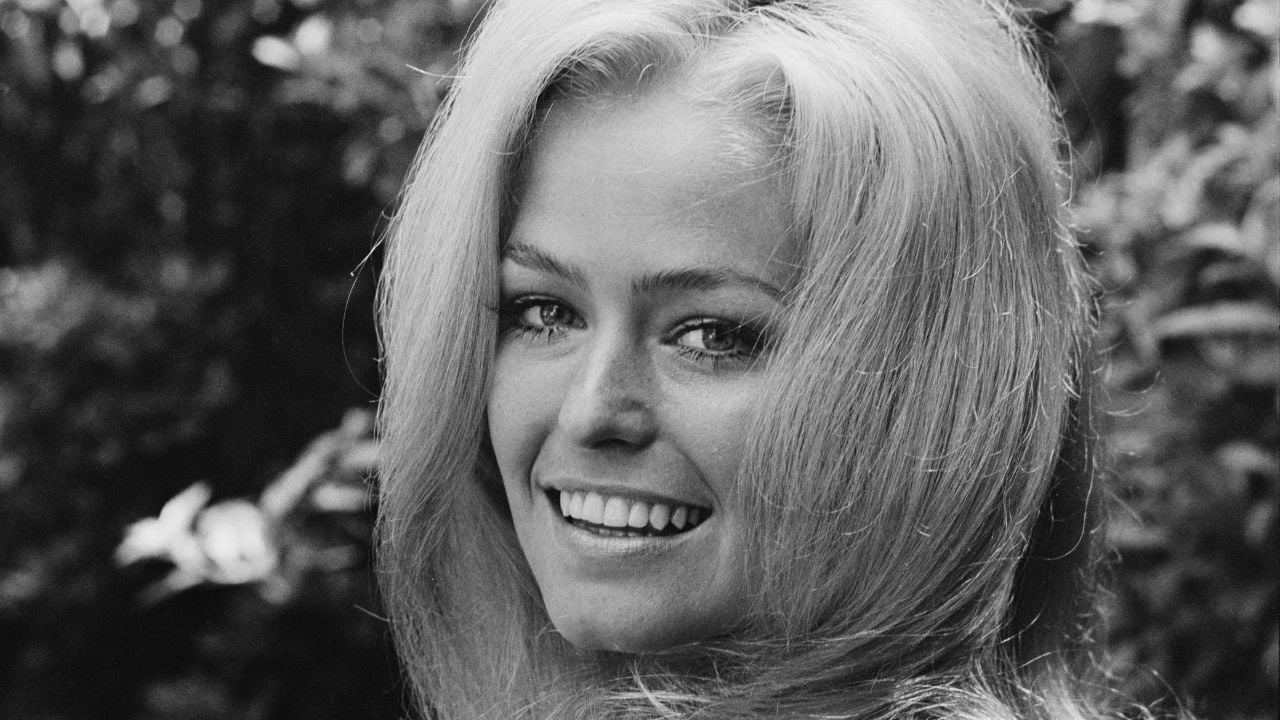 A woman with long blonde hair smiles at the camera in an outdoor setting, with blurred greenery in the background. The photo is black and white.