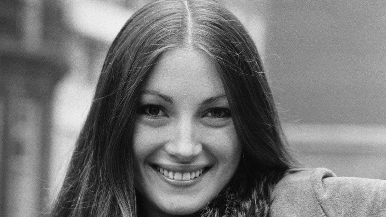 A young woman with long, straight hair smiles warmly at the camera in a black and white photo. She wears a coat and is outdoors, with a blurred background.