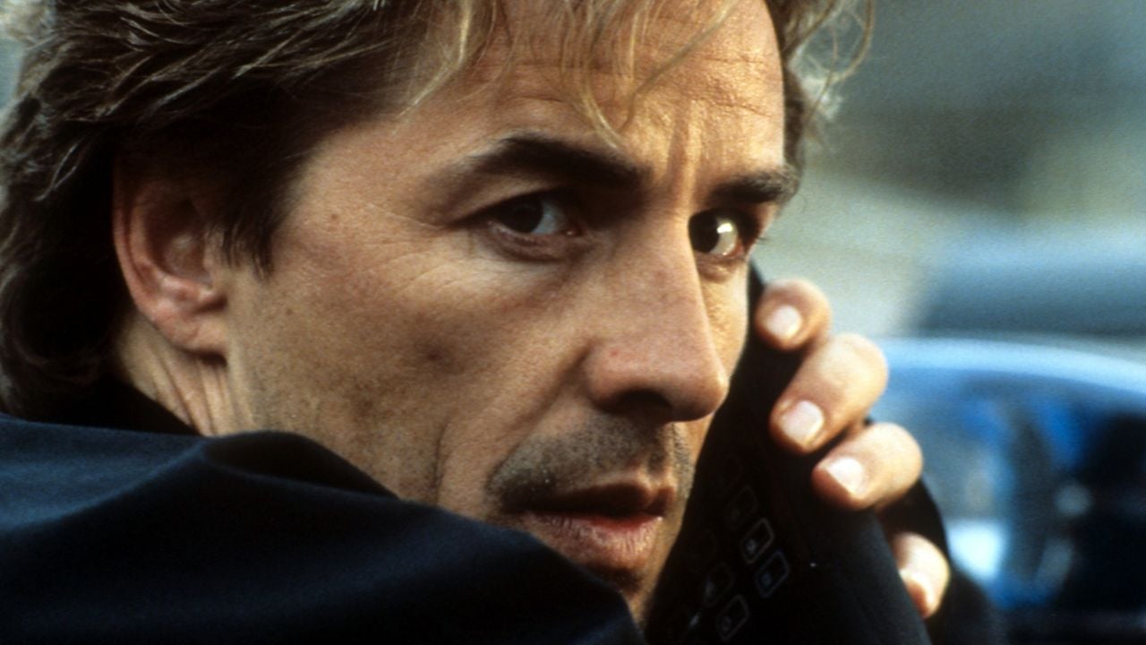 A man with tousled hair looks over his shoulder while holding a black phone to his ear, appearing tense or cautious.