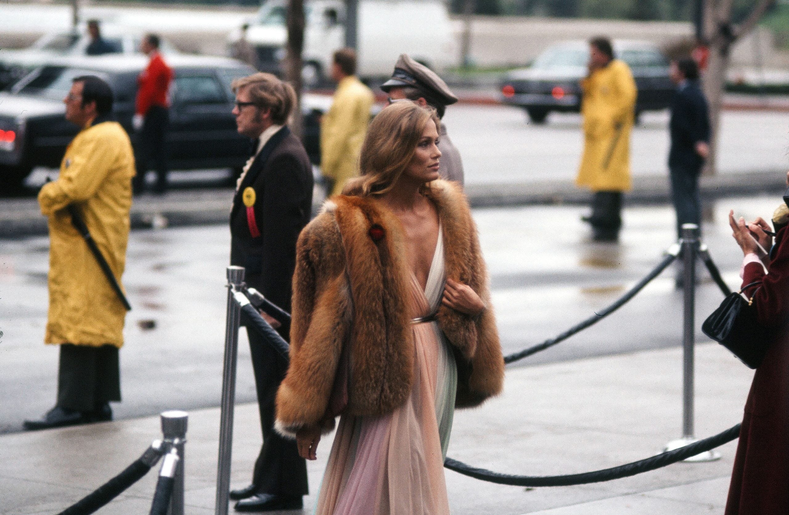 A woman in a fur coat and elegant dress walks past a roped-off area, with people in yellow raincoats and others standing nearby on a wet sidewalk.