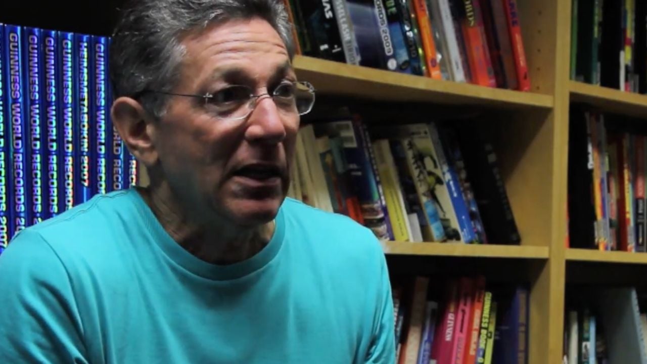 A man wearing glasses and a turquoise shirt is sitting and speaking in front of bookshelves filled with various colorful books.