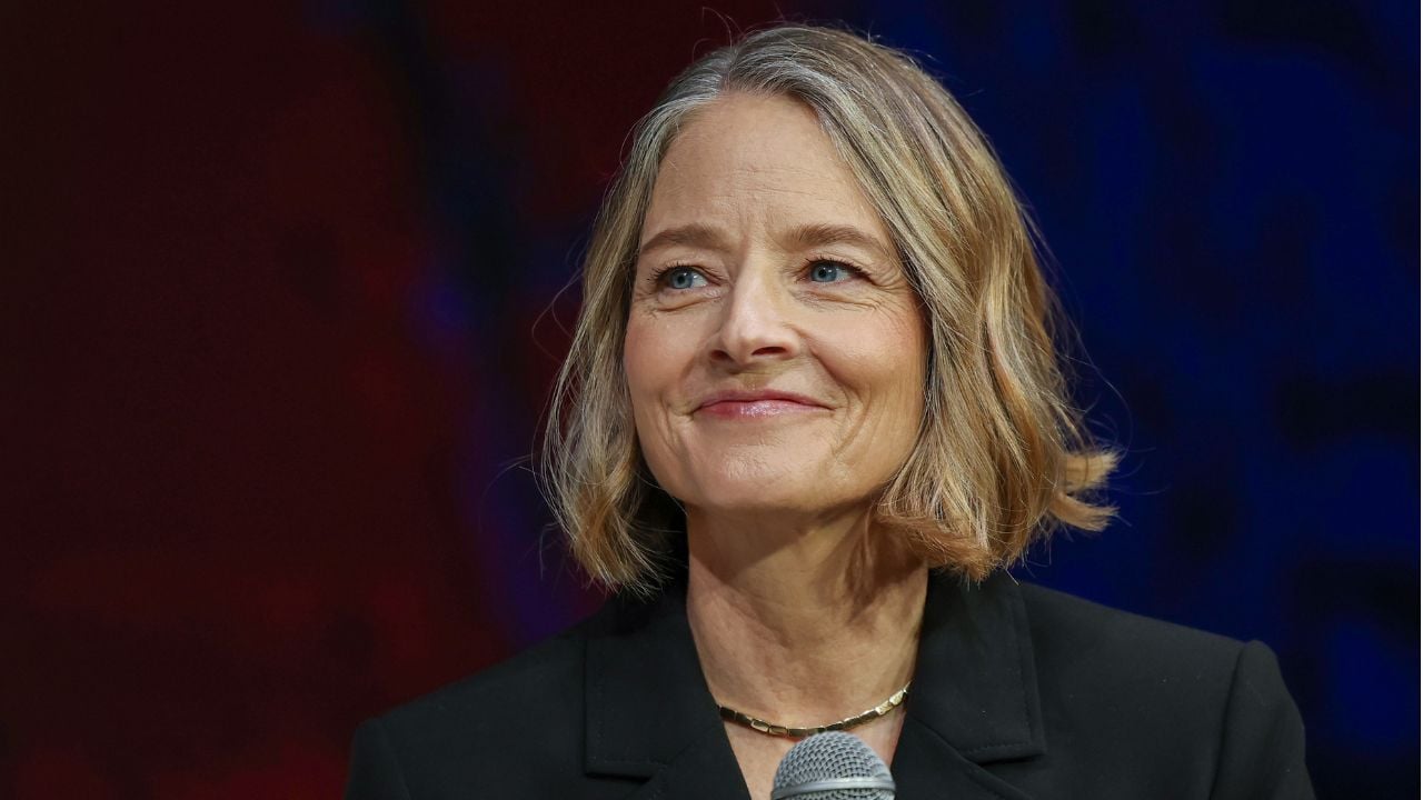 A woman with short, blonde hair wearing a black top and gold necklace holds a microphone and smiles while looking to the side against a dark, blurred background.