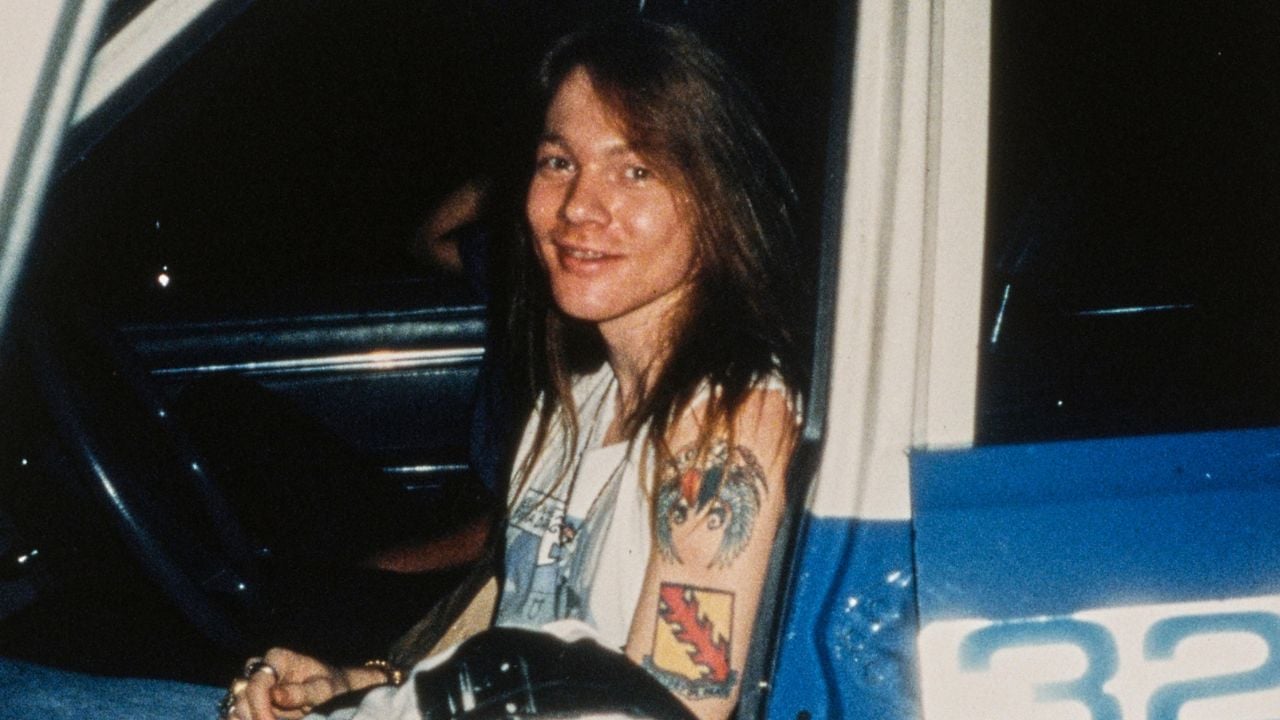 Smiling person with long hair and tattoos on their arm sits in the open door of a blue and white vehicle, wearing a sleeveless shirt.