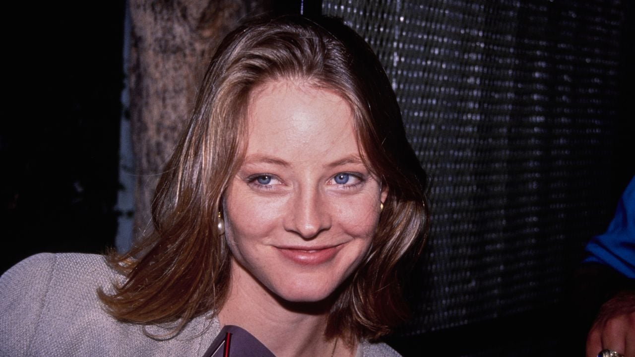 A woman with shoulder-length light brown hair and blue eyes smiles slightly, wearing a light-colored blazer. The background is dark and blurred, with a tree and some mesh fencing visible.
