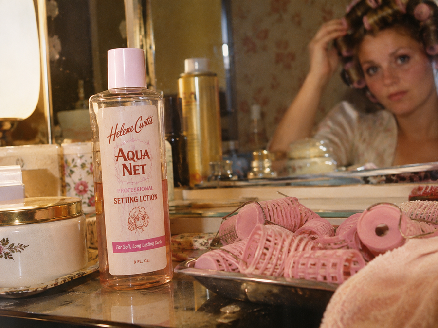 A bottle of Aqua Net setting lotion on a vanity, surrounded by hair rollers and beauty items. In the mirror, a woman in curlers is touching her hair, getting ready.