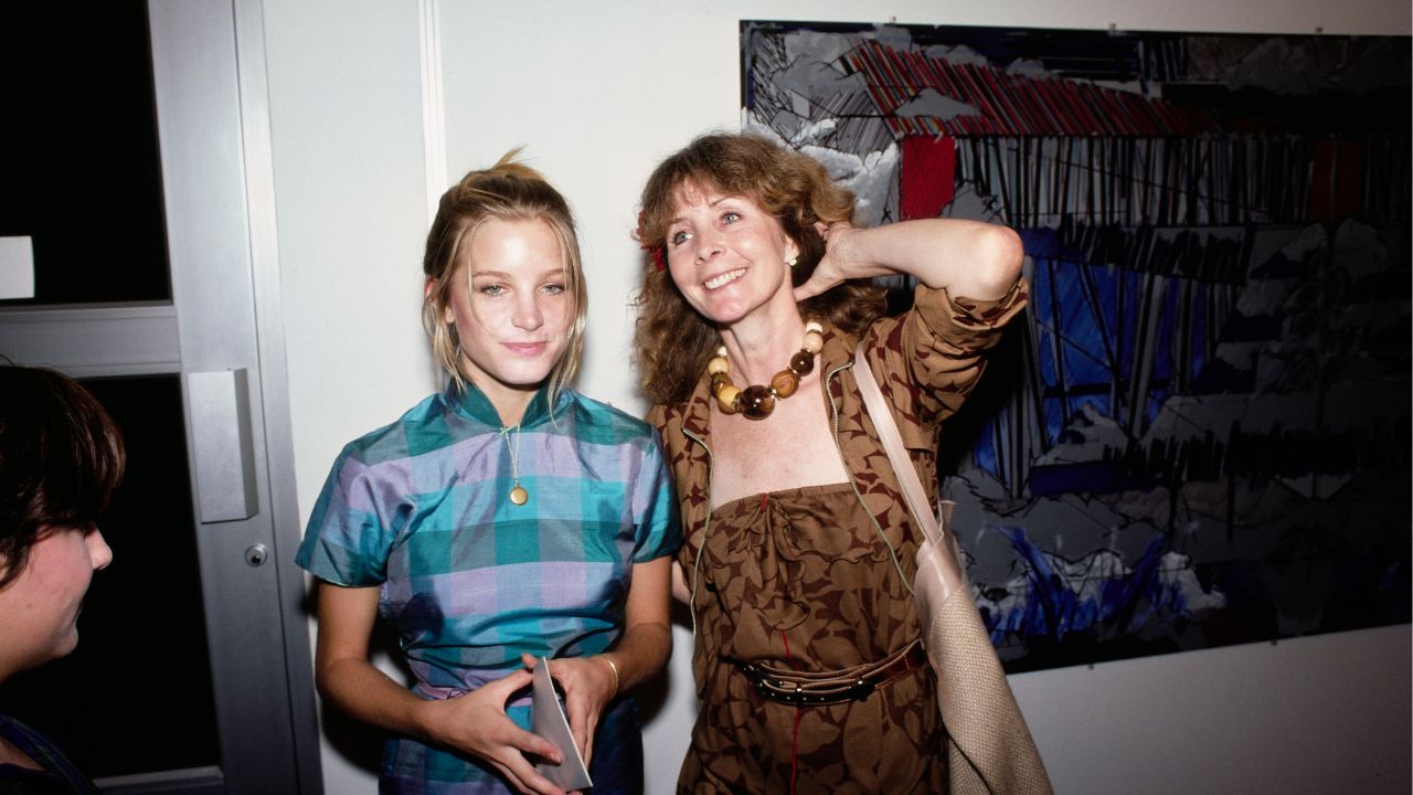 Two women stand together indoors, smiling. One wears a blue plaid shirt, the other a brown patterned dress with a chunky necklace. An abstract painting hangs on the wall behind them.