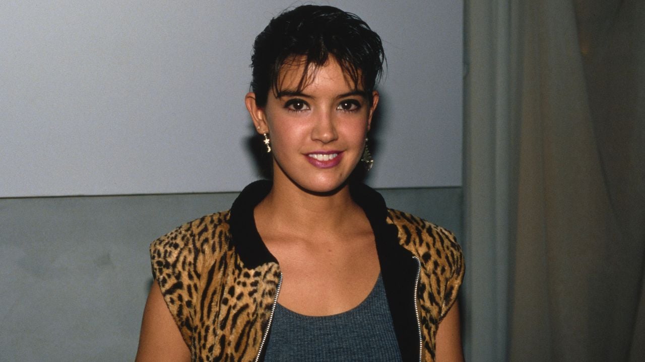 A young woman with short dark hair wears a sleeveless, leopard-print jacket over a gray top. She is smiling and standing indoors against a plain background.