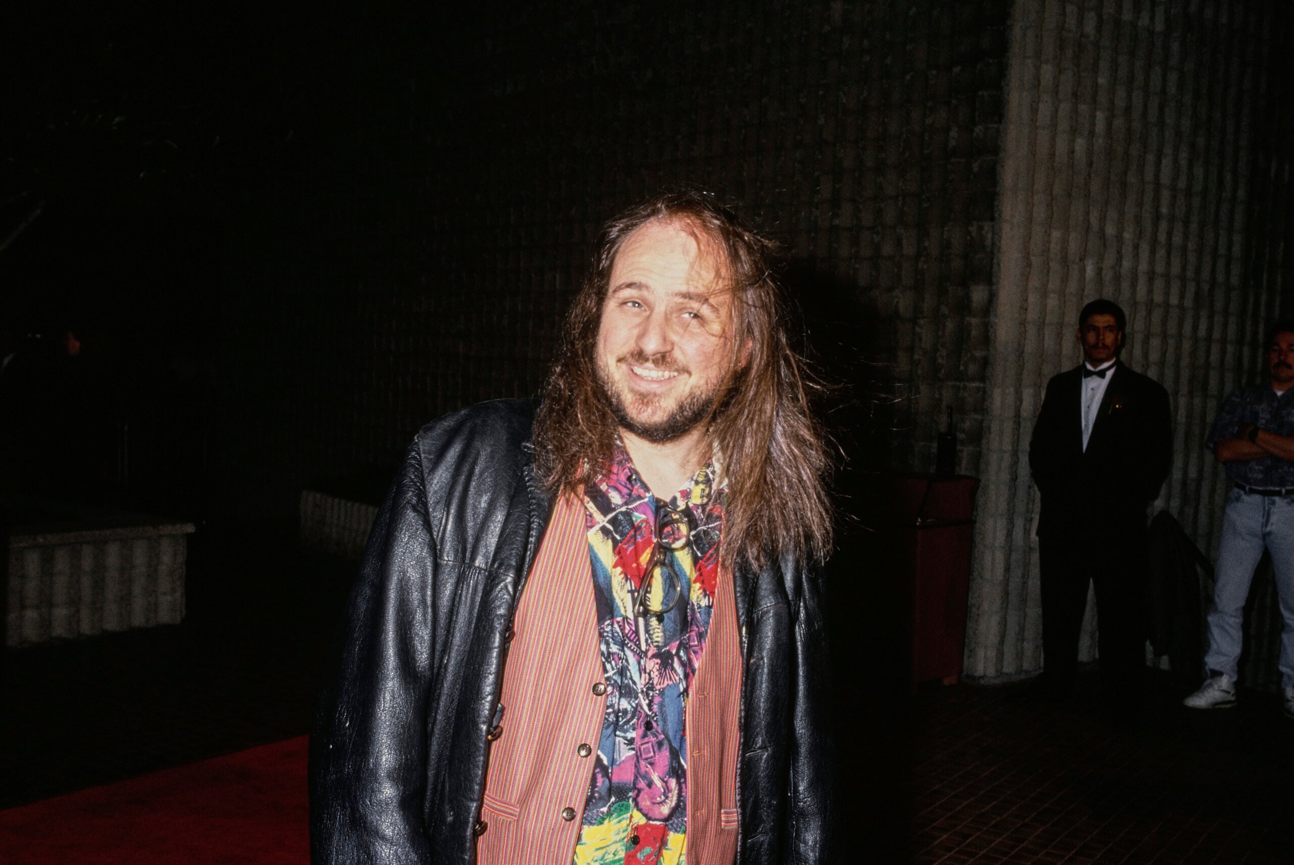 A man with long hair and a beard smiles while wearing a colorful shirt and leather jacket, standing on a red carpet with people in the background at night.