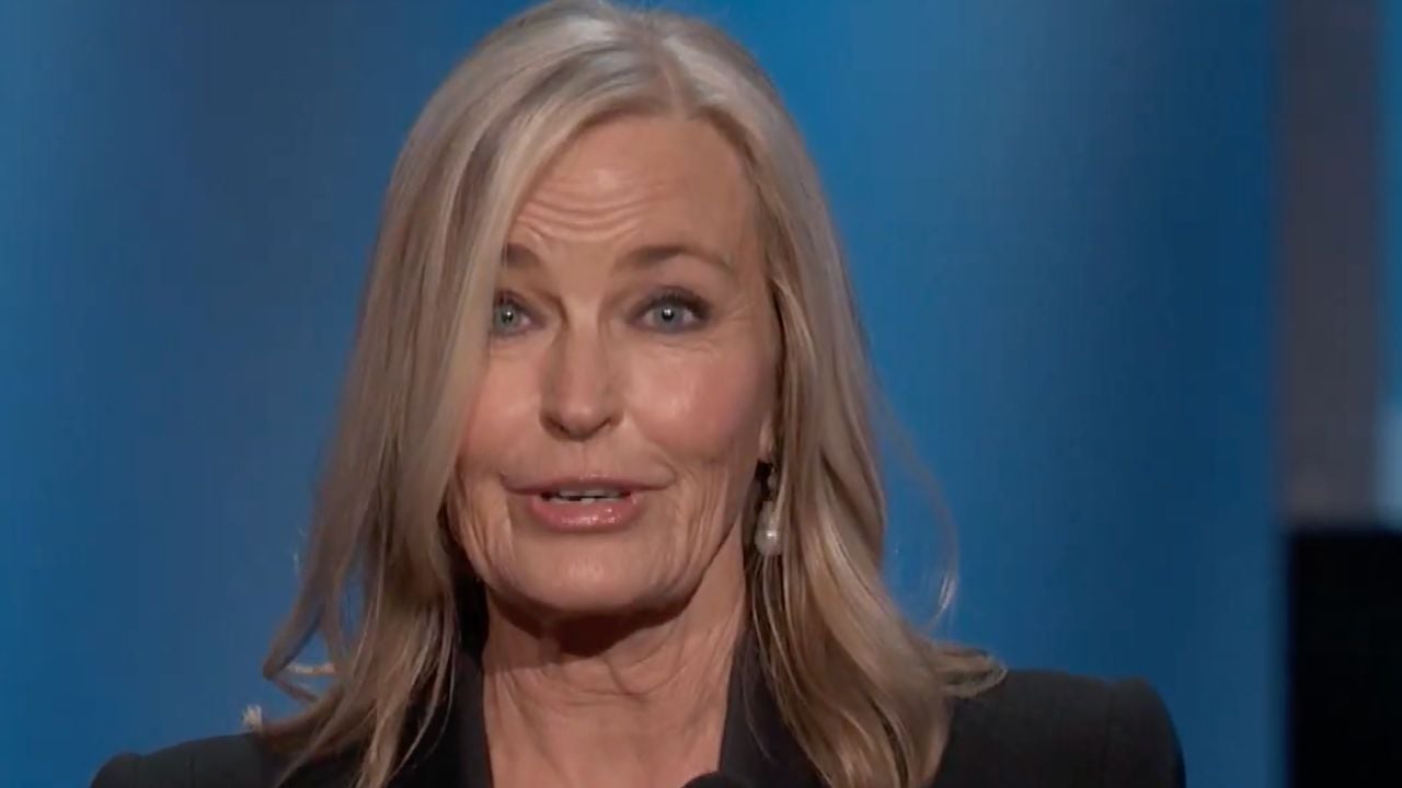 A woman with long blonde hair and light skin speaks in front of a blue background, wearing a black outfit and pearl earring.