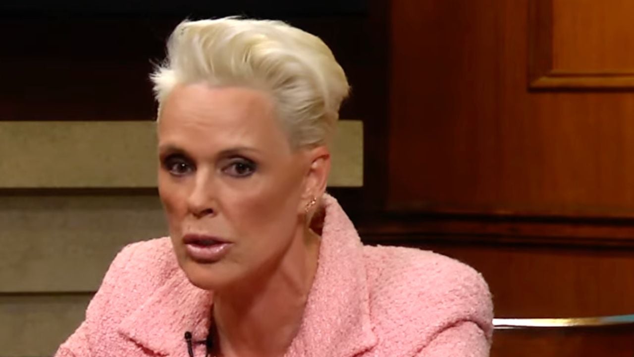A person with short, slicked-back platinum blonde hair wearing a textured pink blazer is speaking in a studio setting.