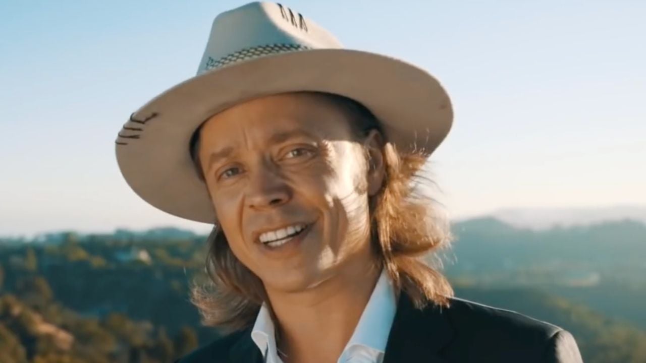 A person wearing a light-colored wide-brimmed hat and a suit jacket smiles outdoors, with hills and a clear sky in the background.