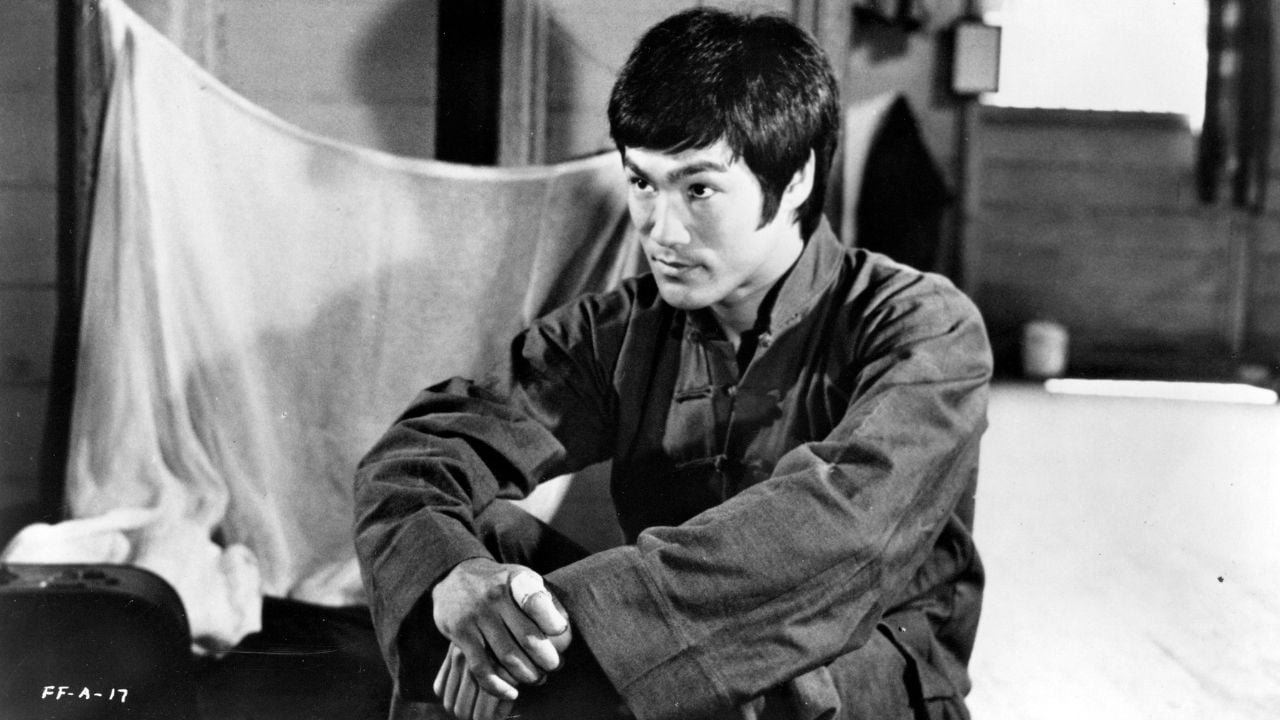 A man in traditional martial arts clothing sits on the floor indoors, resting his arms on his knees and looking to the side with a thoughtful expression.