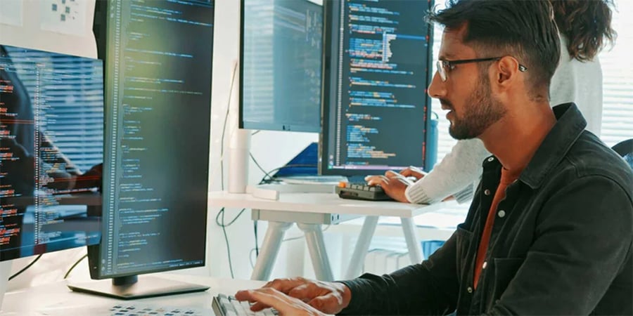 A man sits at a desk typing on a keyboard, looking at two large monitors displaying lines of code. Another person stands in the background working on a laptop. The setting appears to be a modern office.