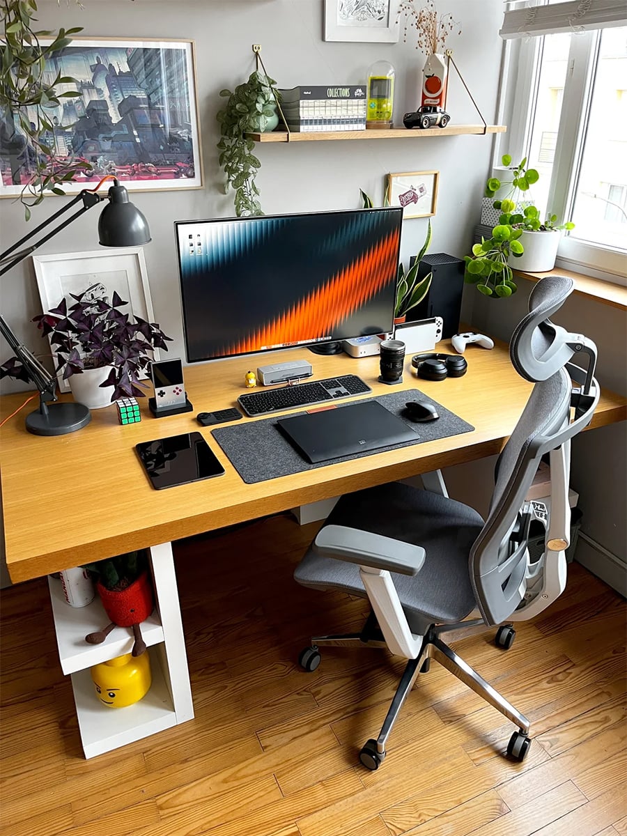 A modern desk setup with a widescreen monitor, keyboard, mouse, tablet, and various gadgets. Plants, books, and framed artwork decorate the space. A gray ergonomic chair is placed at the wooden desk near a window.