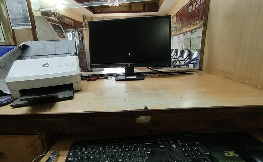 A wooden desk with a computer monitor, a keyboard, and an HP printer; empty chairs and large windows are visible in the background.