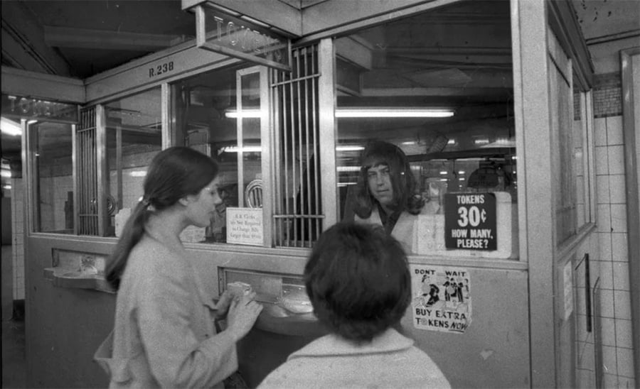 A subway token booth attendant serves two women. Signs on the window show tokens cost 30¢. The scene is black and white, set in an older subway station.