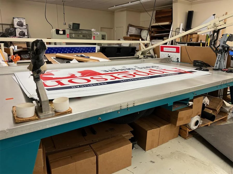 Large “Henderson” campaign sign on a worktable in a cluttered print shop, surrounded by boxes, tools, and printing equipment.