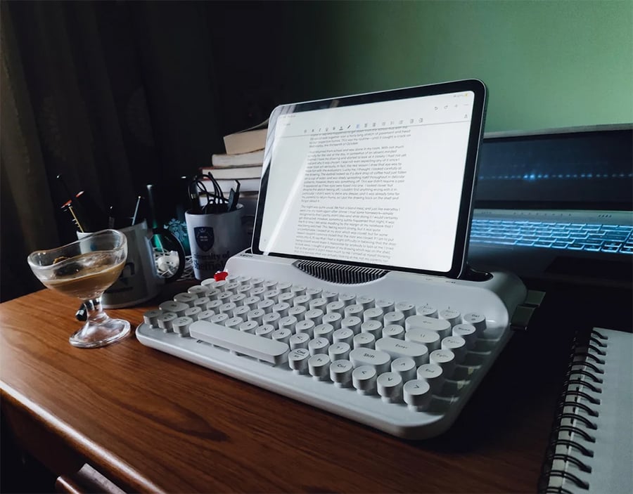A tablet is placed on a typewriter-style keyboard on a wooden desk, with a cup, books, and a notebook nearby. The tablet displays a document in a word processor.