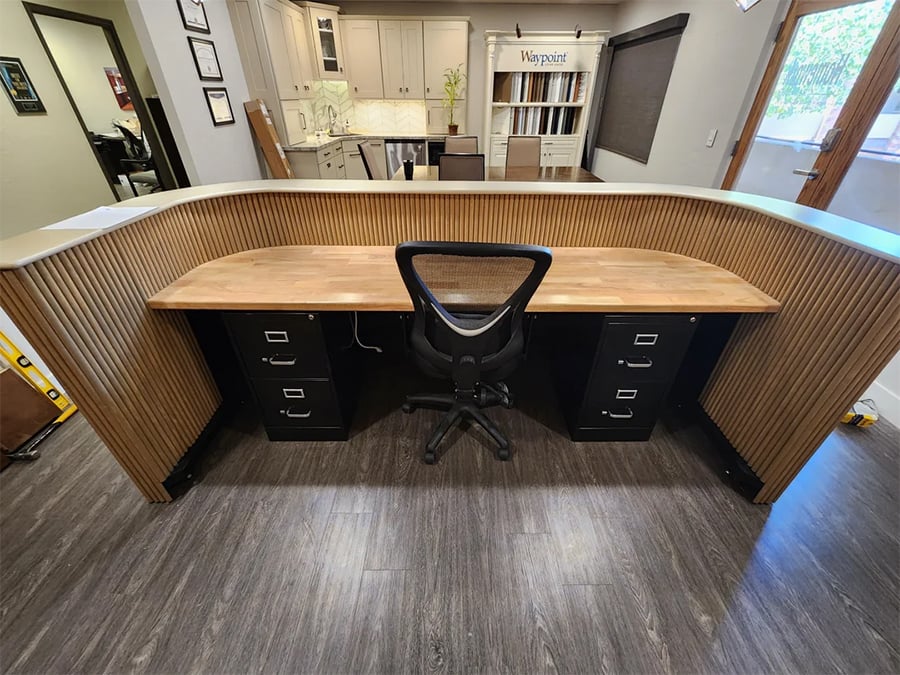 A modern reception desk with a wooden countertop, curved wood paneling, a black office chair, and two black filing cabinets, set on dark flooring in a well-lit office area.
