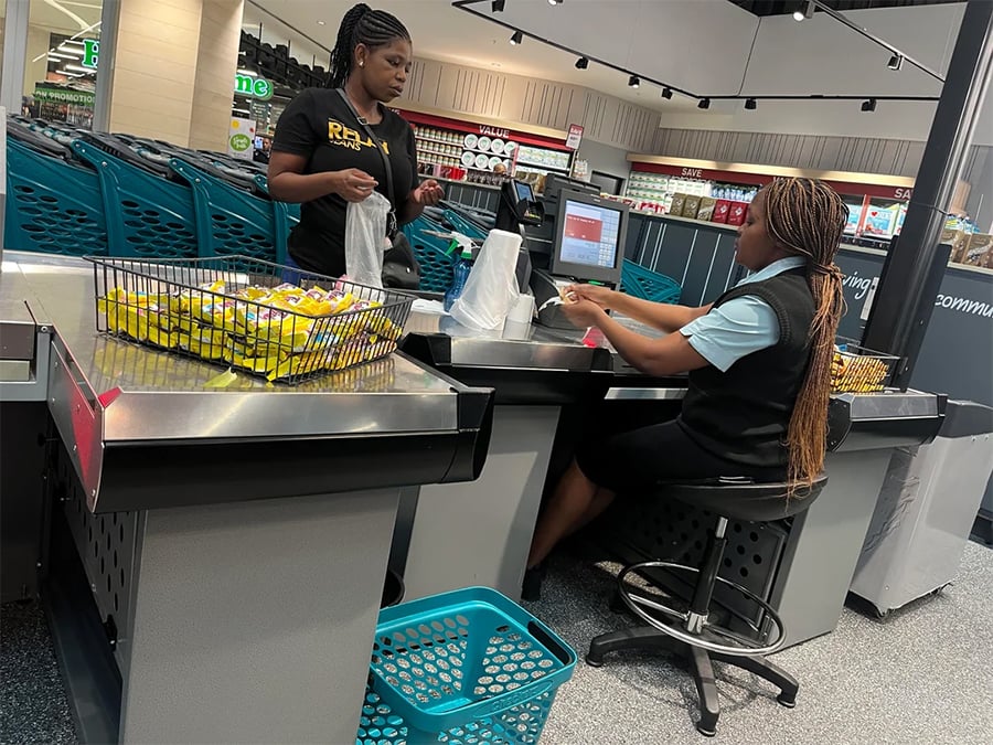 A cashier sits at a checkout counter scanning items as a customer places groceries into bags. Blue shopping baskets and store shelves are visible in the background.