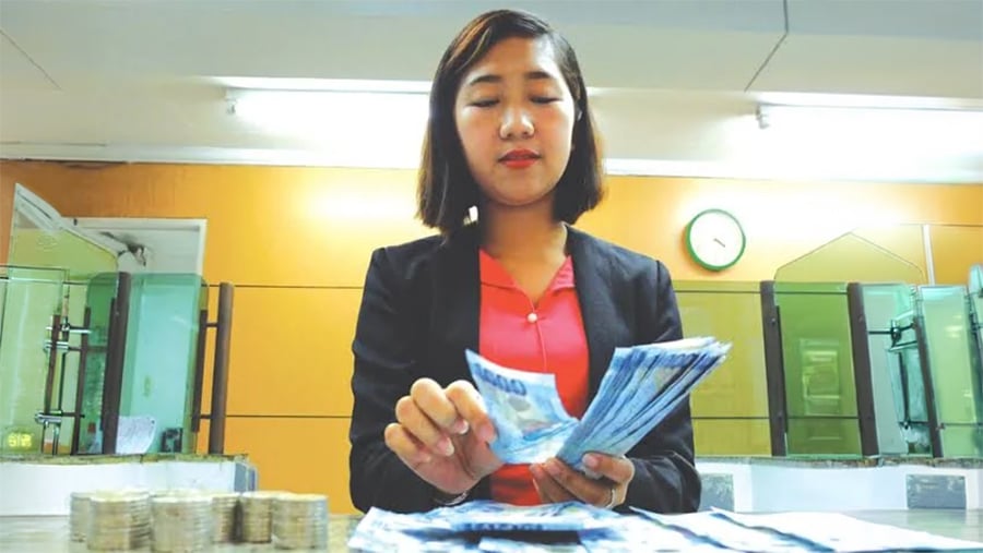 A woman in business attire counts a stack of paper bills at a counter, with stacks of coins beside her and a clock on the wall in the background.