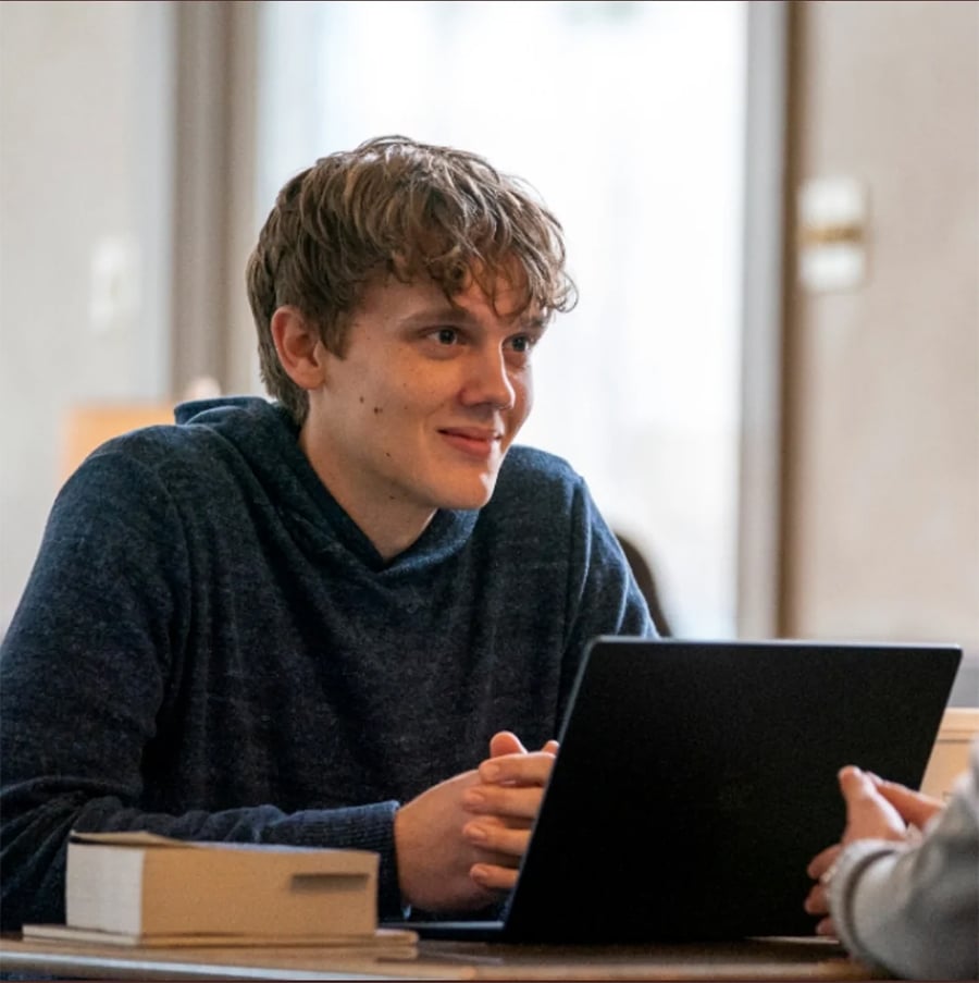 A young person with curly hair sits at a table, smiling and looking at someone off-camera, with an open laptop and a book in front of them.