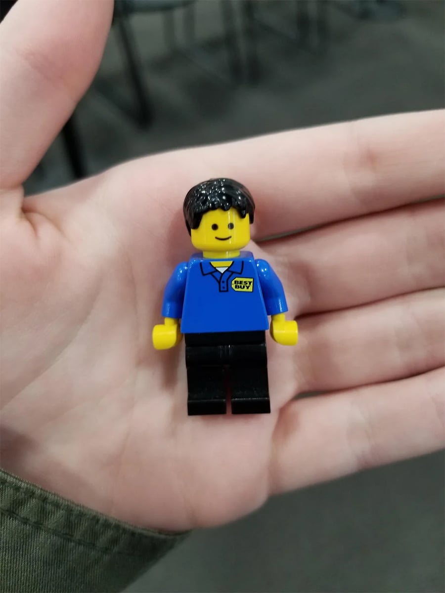 A hand holds a LEGO minifigure wearing a blue shirt with a yellow badge, black pants, and black hair.