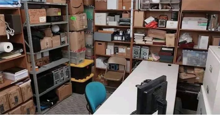 A cluttered storage room with shelves full of boxes, office supplies, and equipment. Two chairs and a computer monitor are next to a white table in the center.